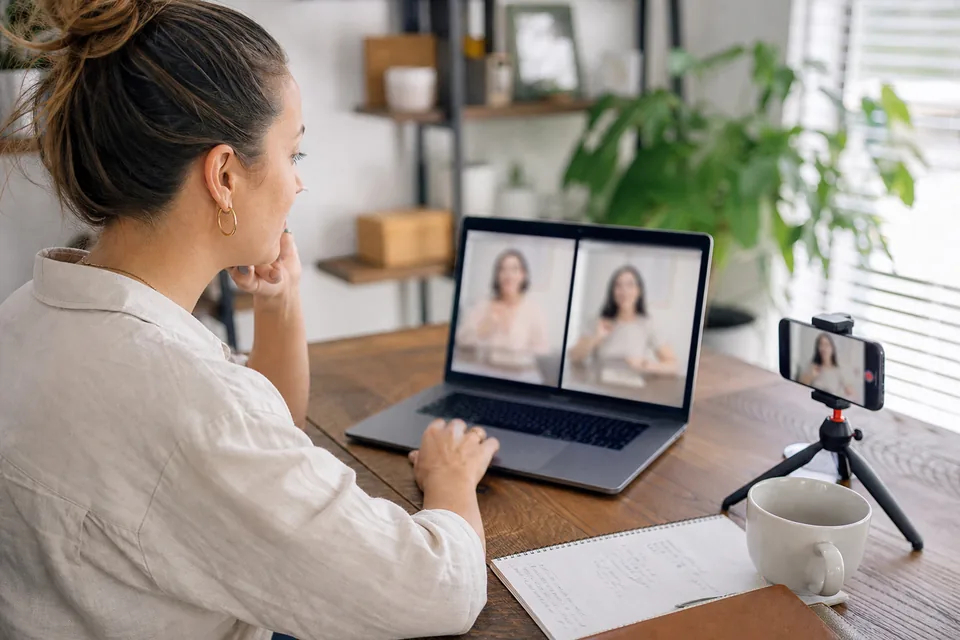 Marketer at a home office desk reviewing two talking-presenter video ad drafts side by side on a laptop, phone on a small tripod beside it