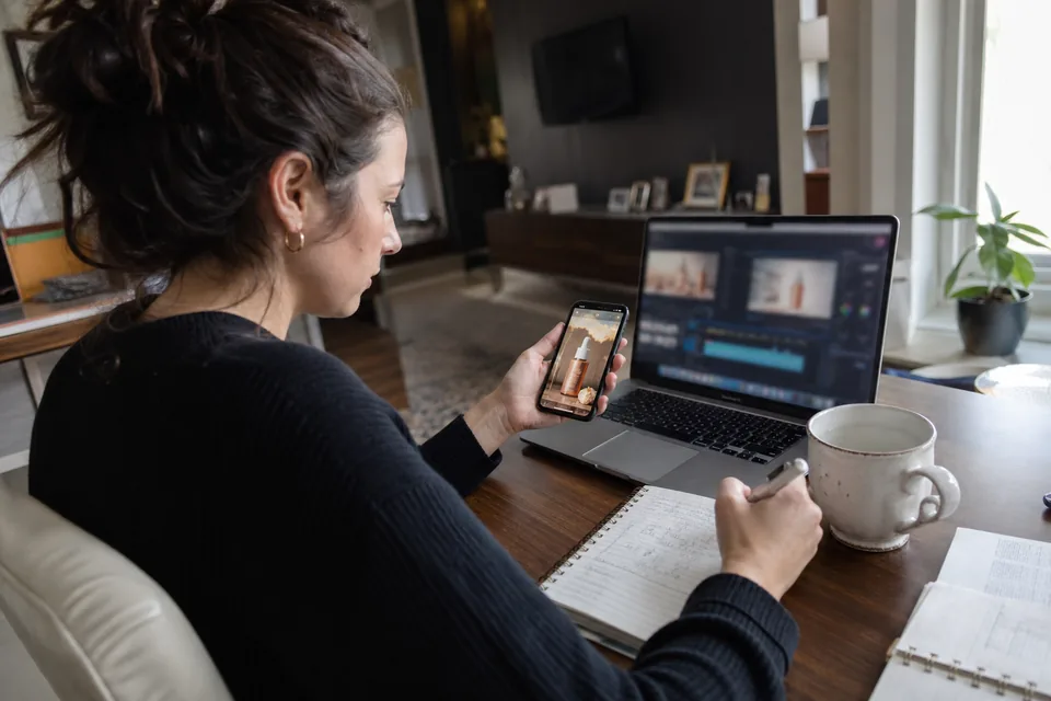 Performance marketer at a desk comparing two AI video generation dashboards on a laptop while a vertical product ad plays on her phone