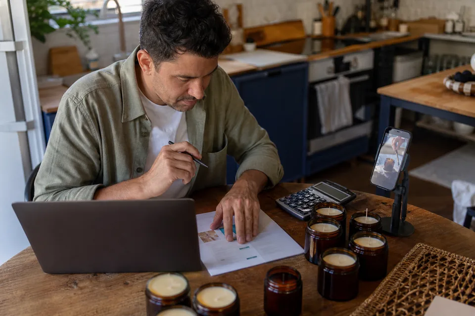Small brand owner reviewing a subscription invoice on a laptop next to product samples and a phone showing a vertical ad