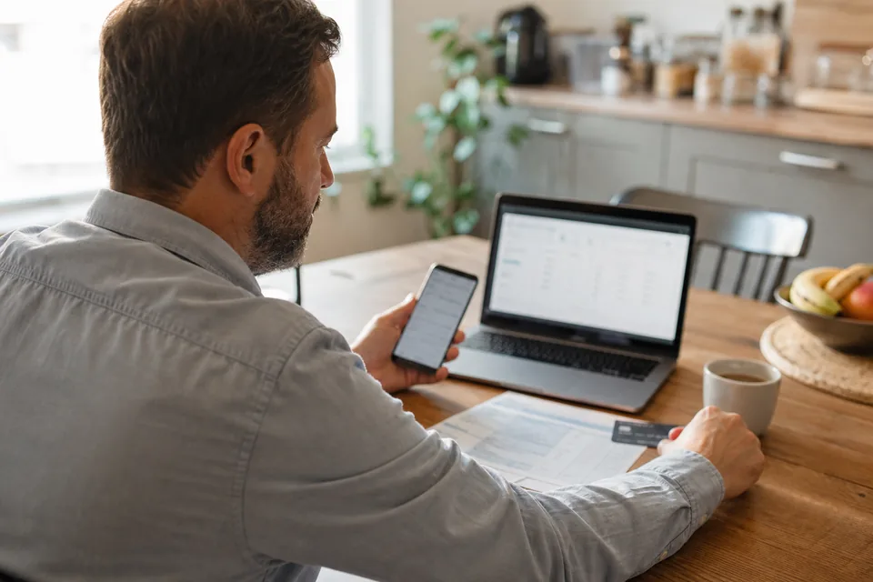 Business owner checking a card statement on a laptop with a phone in hand at a kitchen table